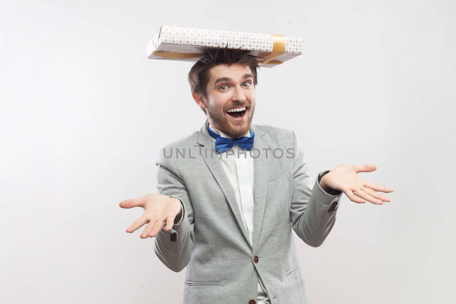 Portrait of joyful bearded man standing with present box on head, expressing happiness, saying take your gift, wearing grey suit and blue bow tie. Indoor studio shot isolated on gray background.