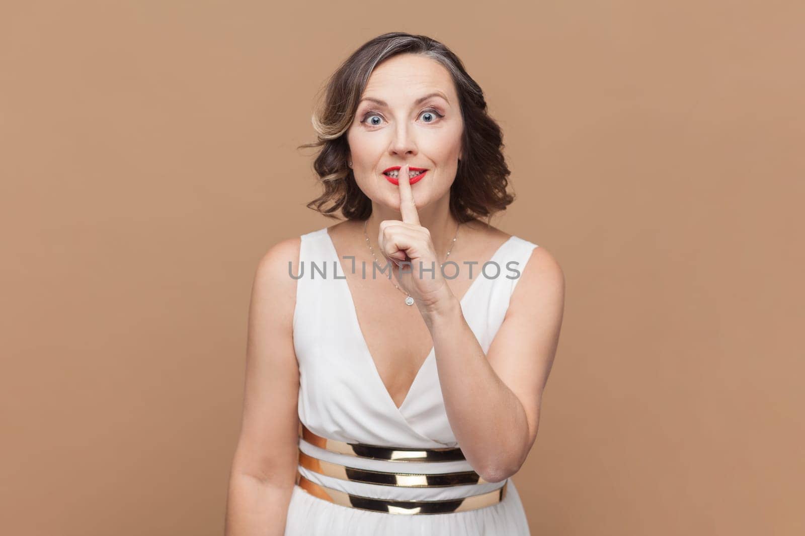 Portrait of woman makes silence or hush gesture, keeps index fingers over lips, tells secret information to someone, wearing white dress. Indoor studio shot isolated on light brown background.