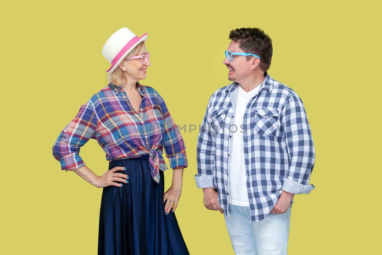 Portrait of adult woman and man in casual checkered shirt and colorful eyeglasses standing together and looking at each other with smile. Indoor studio shot isolated, on yellow background