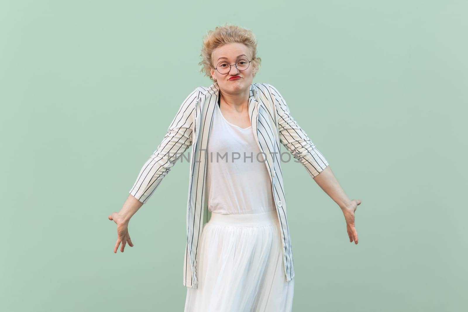 Portrait of confused puzzled young adult blonde woman wearing striped shirt and skirt, shrugging shoulders, doesn't know answer and what to do. Indoor studio shot isolated on light green background.