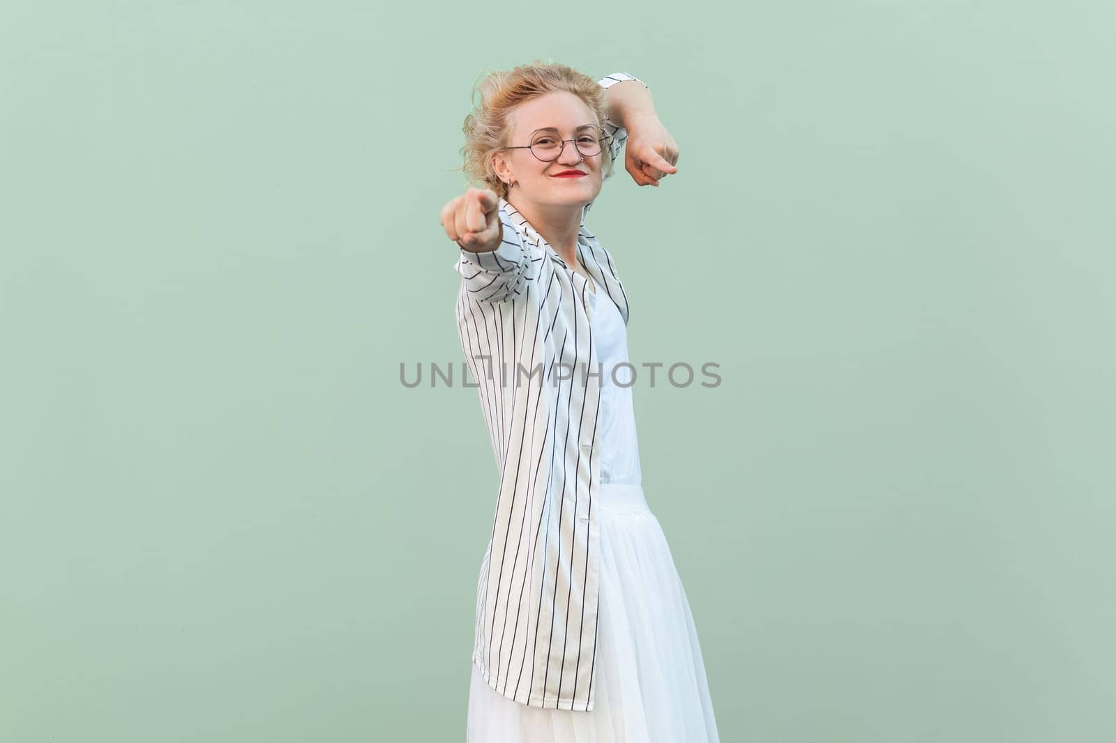 Portrait of joyful cheerful positive blonde woman wearing striped shirt and skirt, pointing index finger to camera, choosing you. Indoor studio shot isolated on light green background.
