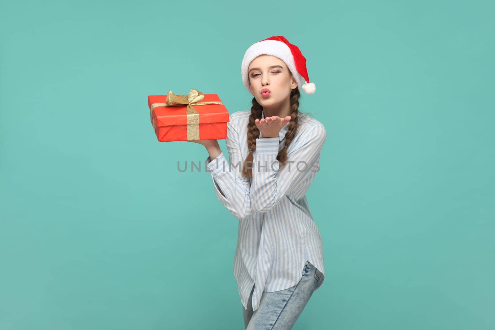 Portrait of charming cute romantic teenager girl with braids wearing striped shirt and Santa Claus hat, holding gift box, sending air kiss. Indoor studio shot isolated on green background.