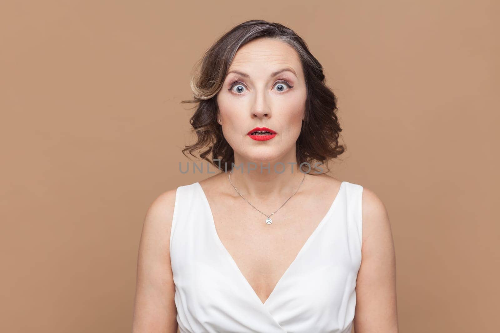 Portrait of shocked astonished middle aged woman with wavy hair looking at camera with big eyes, sees something excited, wearing white dress. Indoor studio shot isolated on light brown background.