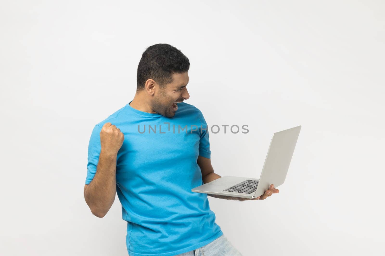 Joyful cheerful unshaven man in blue T- shirt standing holding laptop in hands, clenches fist, celebrating success, finishing project till deadline. Indoor studio shot isolated on gray background.