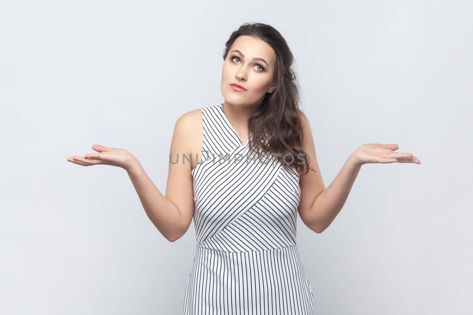 Portrait of brunette woman spreads palms, shrugs shoulders with perplexed expression, being indecisive, has no idea what happened, wearing striped dress. Indoor studio shot isolated on gray background