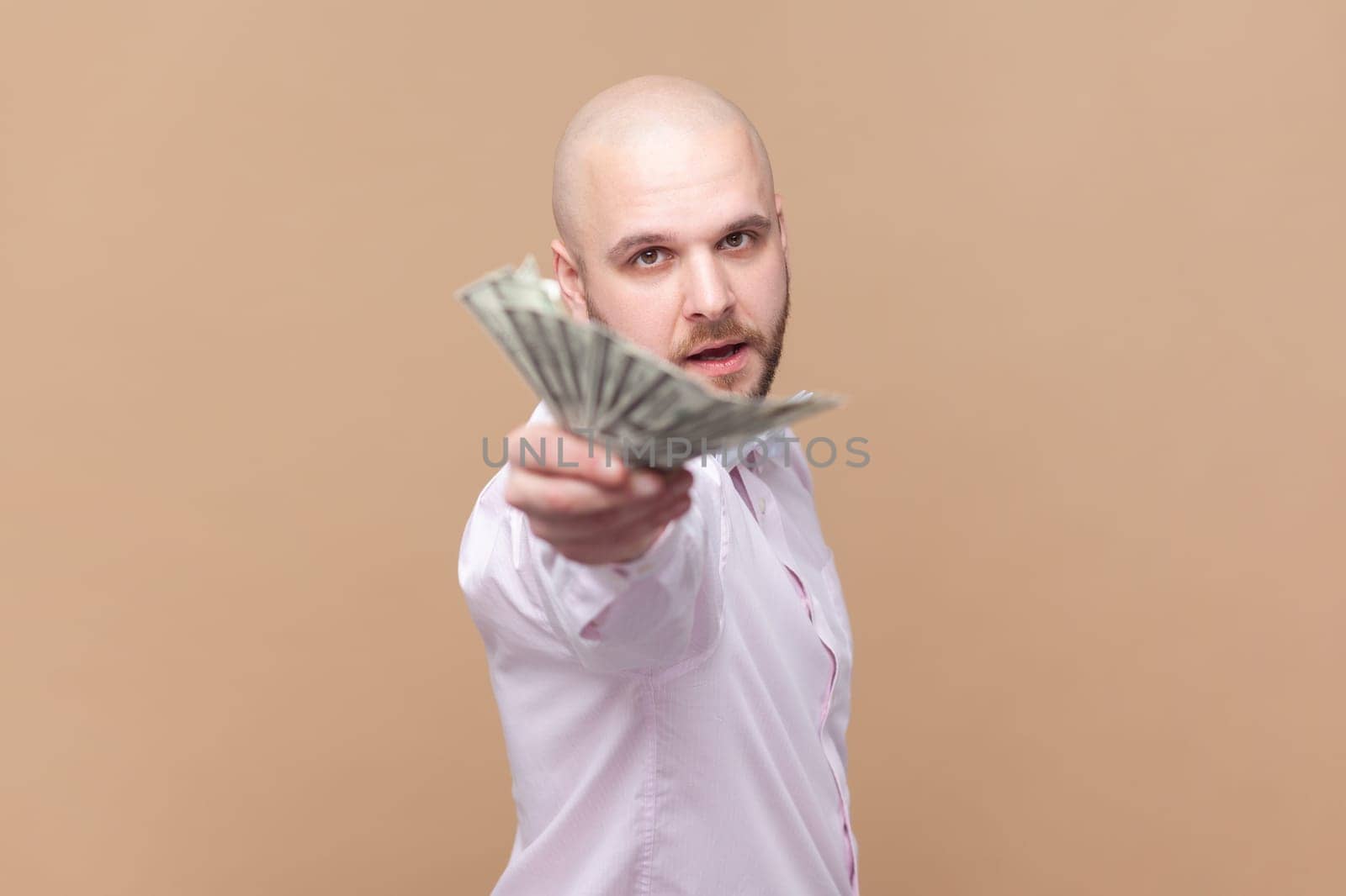 Portrait of handsome rich bald bearded man holding out dollar banknotes, looking at camera, giving money to you, wearing light pink shirt and bow tie. Indoor studio shot isolated on brown background.
