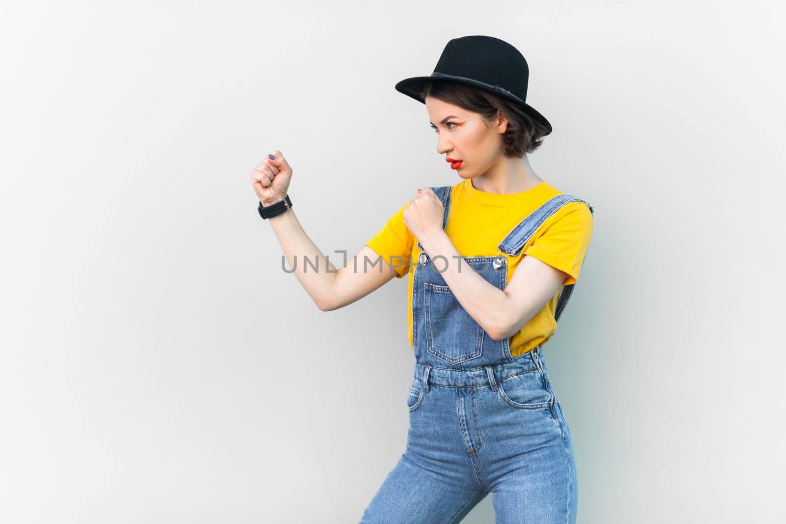 Side view portrait of aggressive hipster woman in blue denim overalls, yellow T-shirt and black hat, keeps fists clenched, being ready to fight. Indoor studio shot isolated on gray background.