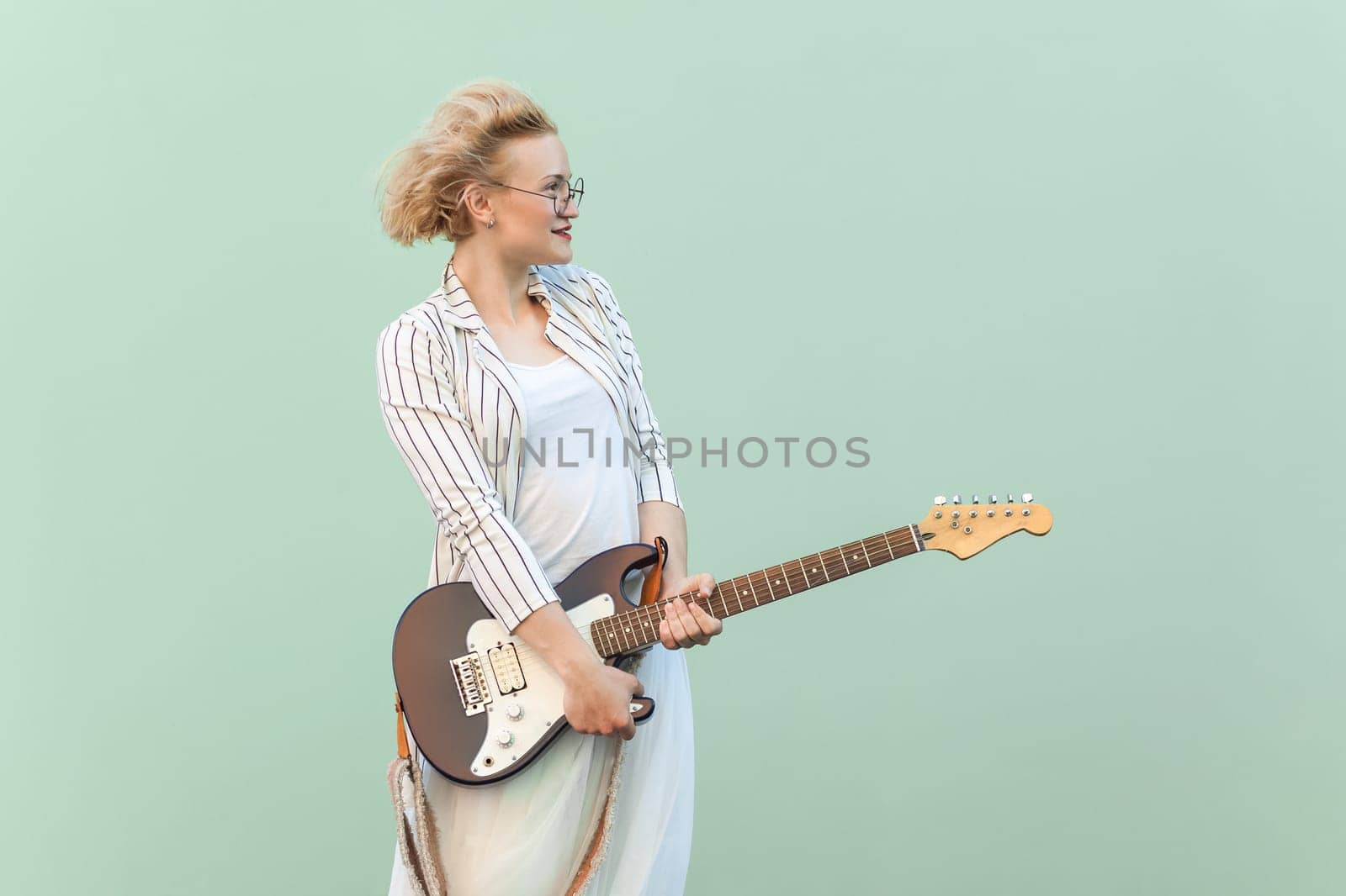 Portrait of beautiful positive blonde woman in white shirt, skirt, and striped blouse with eyeglasses, holding guitar and looking away. Indoor studio shot isolated on light green background.