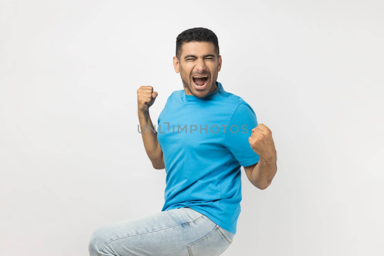 Portrait of unshaven man wearing blue T- shirt standing clenches fists, rejoices his victory and triumph, expresses positiveness, feels like champion. Indoor studio shot isolated on gray background.