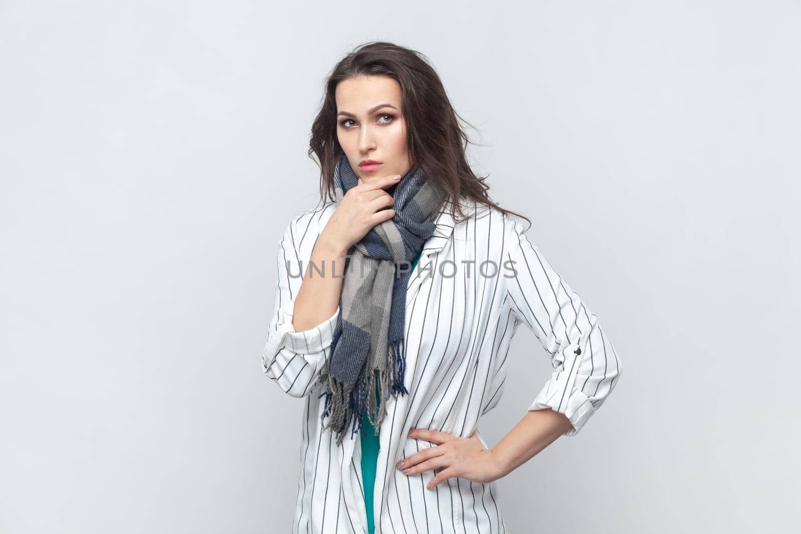 Portrait of pensive thoughtful brunette woman holding her chin, pondering, keeps hand on hip, looking at camera, wearing striped jacket and scarf. Indoor studio shot isolated on gray background.
