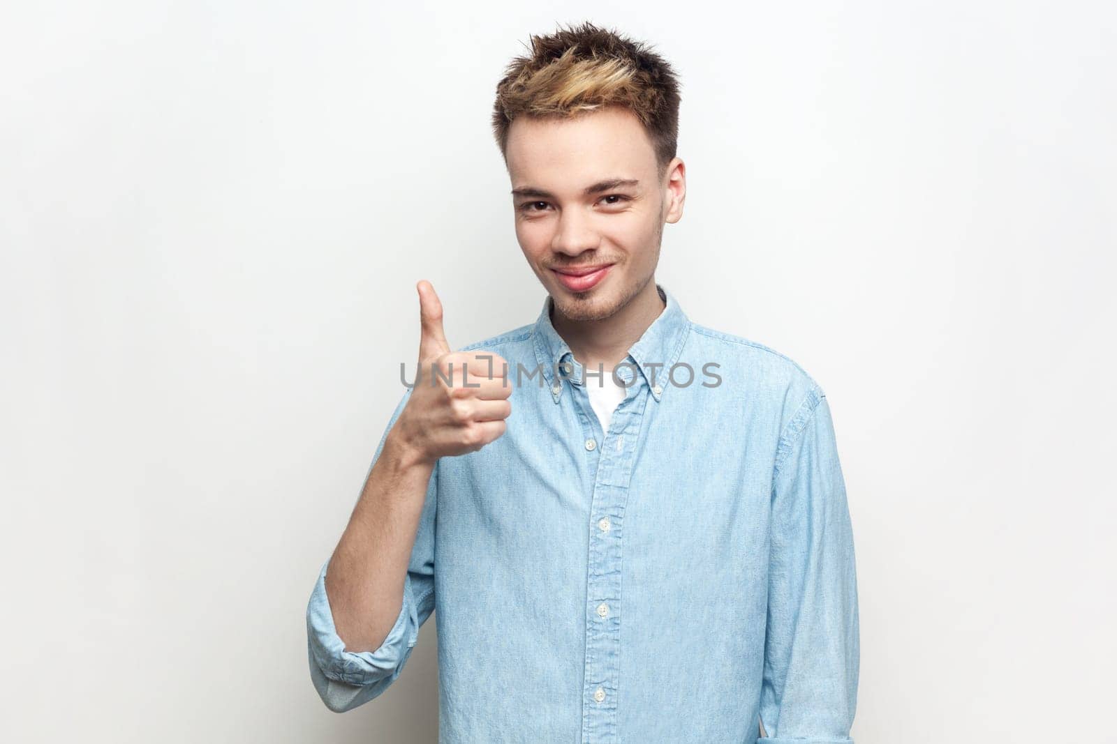 Portrait of smiling happy cheerful man wearing denim shirt showing like gesture, looking at camera, standing with thumb up, expressing happiness. Indoor studio shot isolated on gray background.
