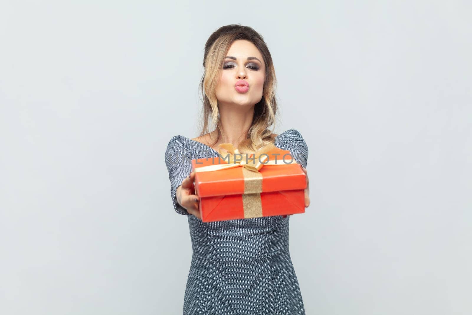 Portrait of positive romantic blonde woman giving present for her husband on anniversary, sending air kiss, wearing grey elegant dress. Indoor studio shot isolated on gray background.