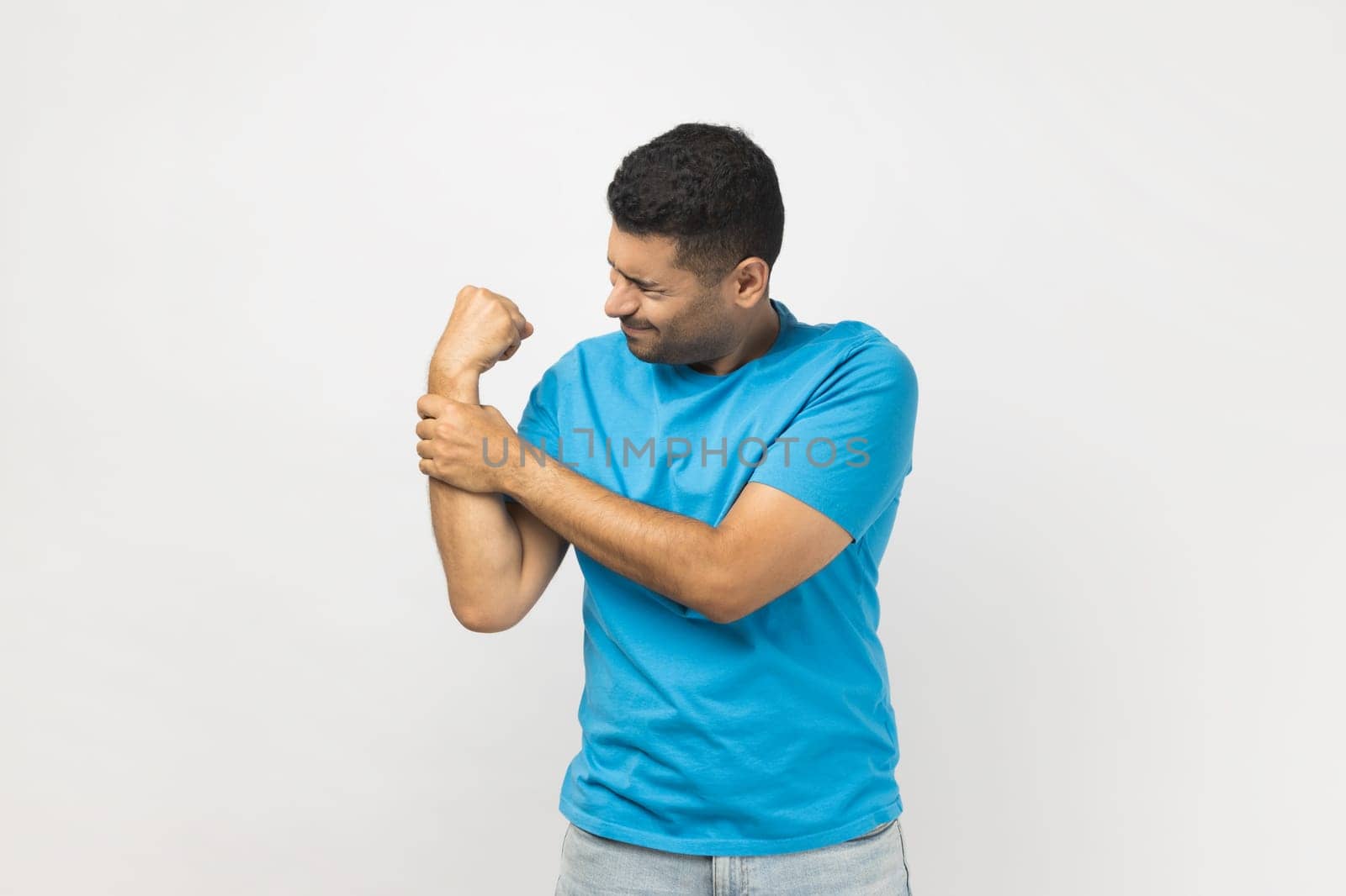 Portrait of unhealthy sick injured unshaven man wearing blue T- shirt standing holding his painful wrist, feels terrible pain, has trauma. Indoor studio shot isolated on gray background.