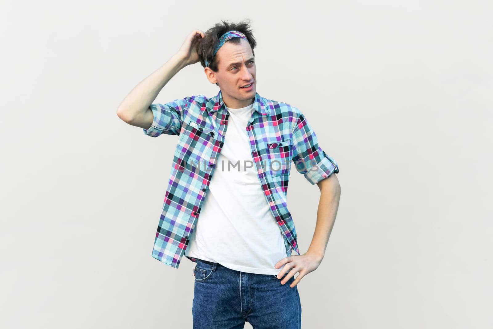 Indignant puzzled man rubbing head, tries to remember necessary information, looks in displeasure, wearing blue checkered shirt and headband. Indoor studio shot isolated on gray background.