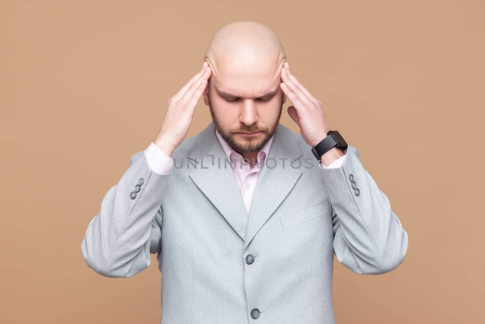 Portrait of sick ill unhealthy bald bearded man standing with hands on head, keeps eyes closed, suffering terrible headache, wearing gray jacket. Indoor studio shot isolated on brown background.