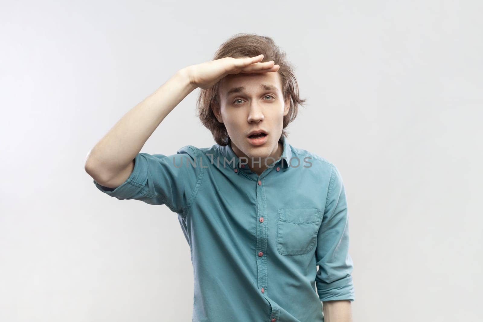 Portrait of concentrated young man looking far away at distance with hand over head, attentively searching for bright future, wearing blue shirt. Indoor studio shot isolated on gray background.