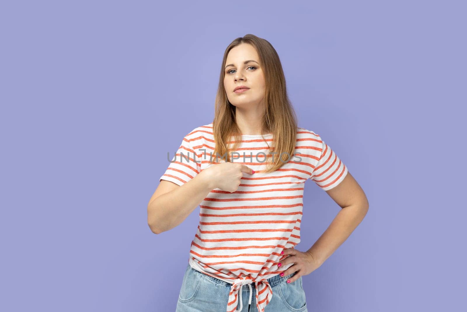 This is me! Arrogant selfish blond woman in striped T-shirt proudly pointing herself, boasting of success, feeling supercilious and self-confident. Indoor studio shot isolated on purple background.