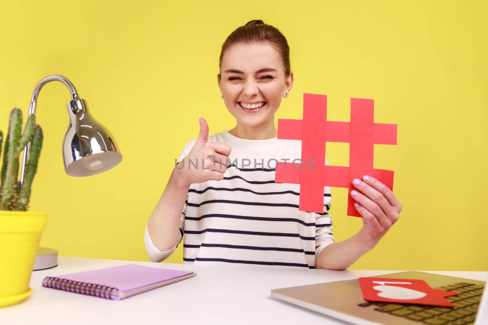 Smiling woman blogger in good mood, showing red hashtag and thumb up like gesture, taking selfie POV, sitting at workplace. Indoor studio studio shot isolated on yellow background.