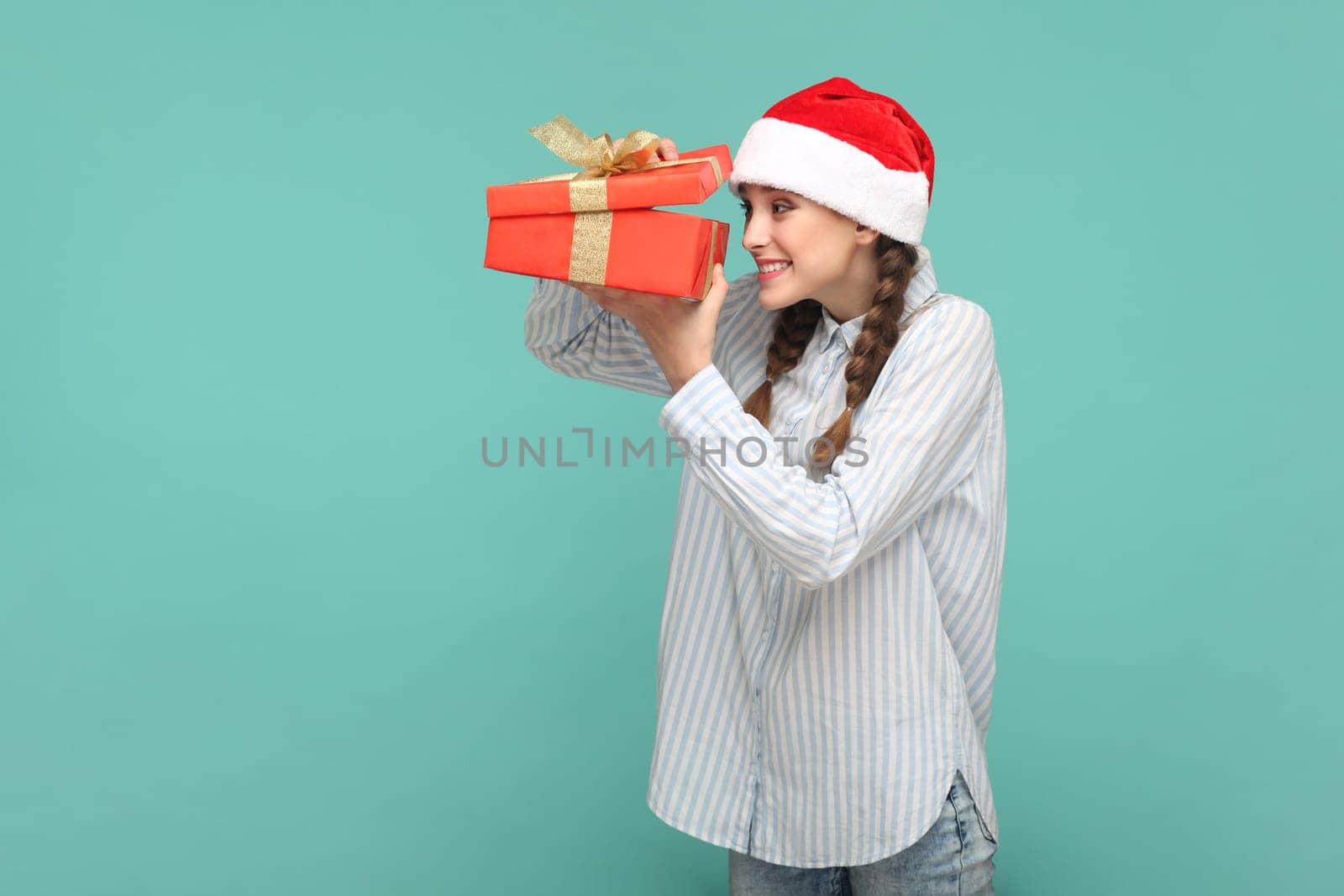Portrait of happy optimistic girl with braids wearing striped shirt and Santa Claus hat, holding present box, looking inside with smile. Indoor studio shot isolated on green background.