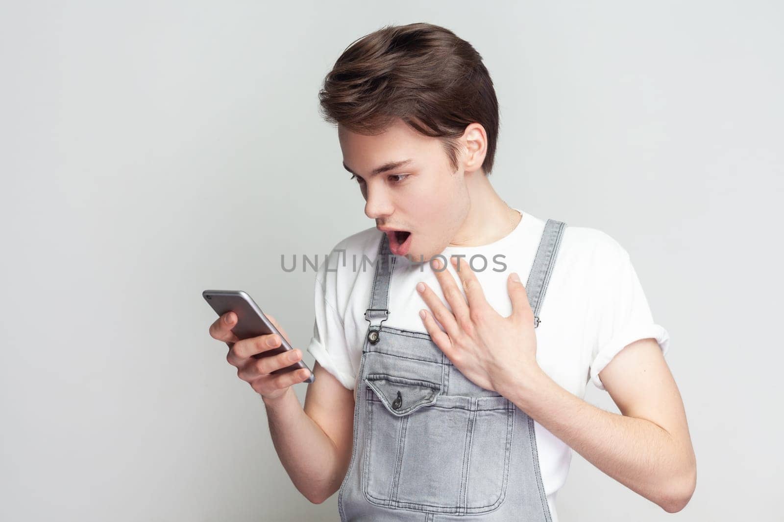 Portrait of shocked astonished amazed young brunette man standing using cell phone, reading shocking news, being surprised, wearing denim overalls. Indoor studio shot isolated on gray background.