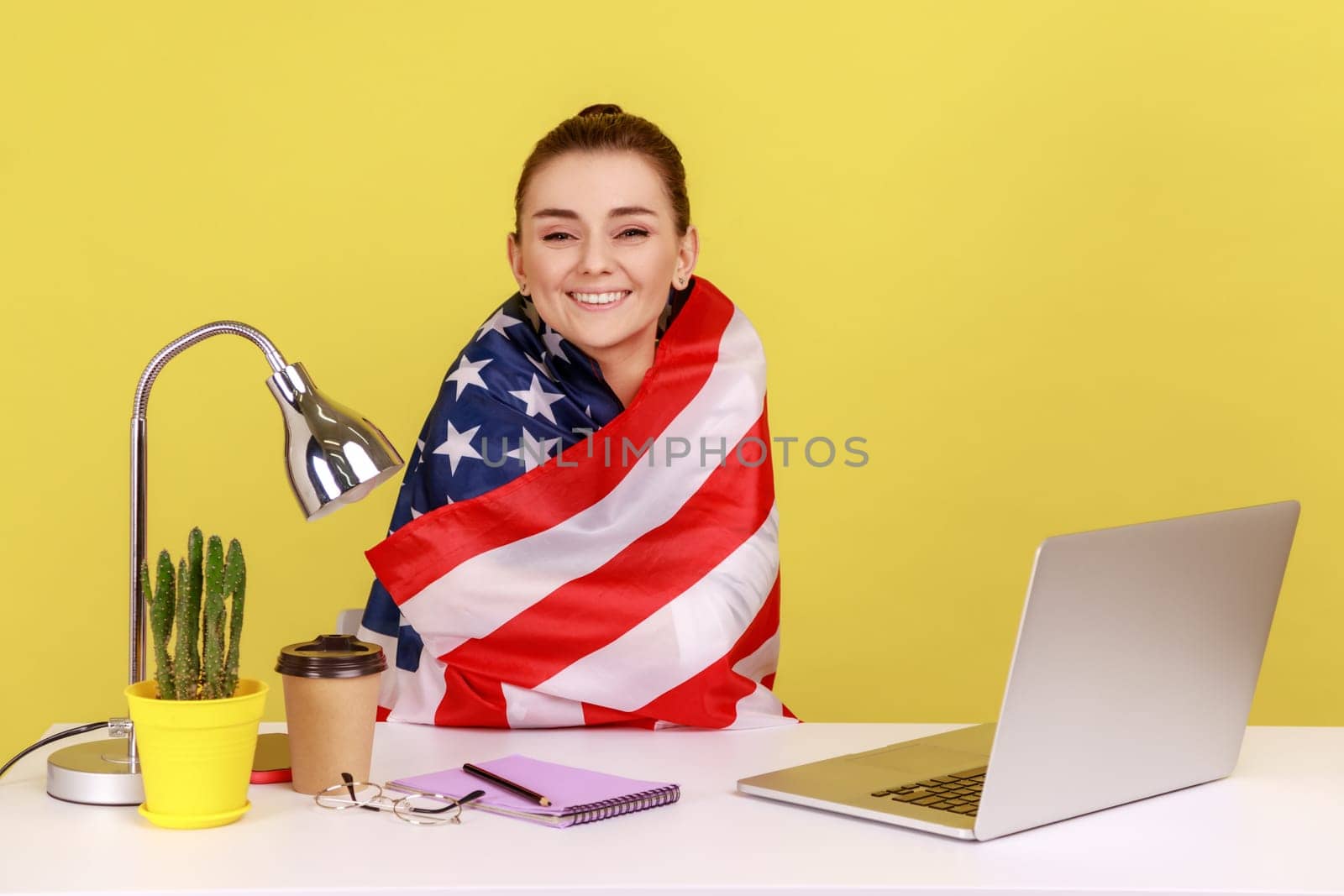 Happy woman sitting wrapped in American flag in office workplace, celebrating labor day or US Independence day 4th of july. Indoor studio studio shot isolated on yellow background.