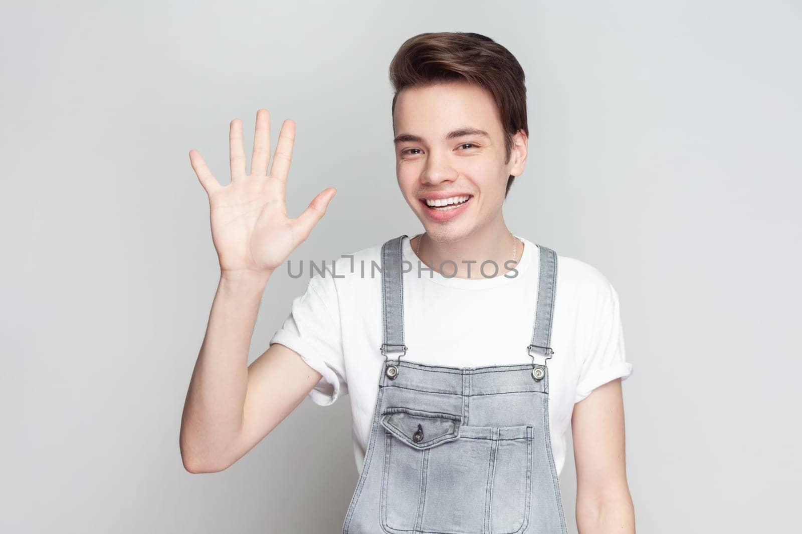 Cheerful friendly young brunette man standing waving palm in hello gesture, meets someone at street, smiles positively, wearing denim overalls. Indoor studio shot isolated on gray background.