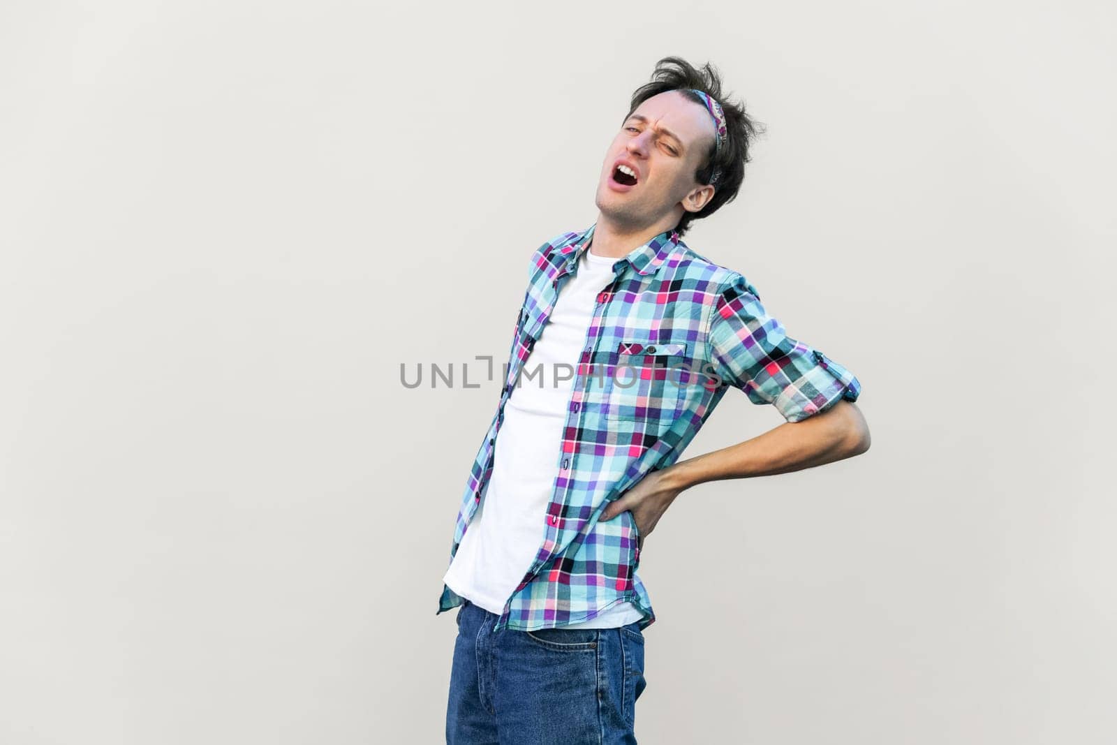 Portrait of man suffers from radiculitis, keeps hand on his painful back, being sick and unhealthy, wearing blue checkered shirt and headband. Indoor studio shot isolated on gray background.