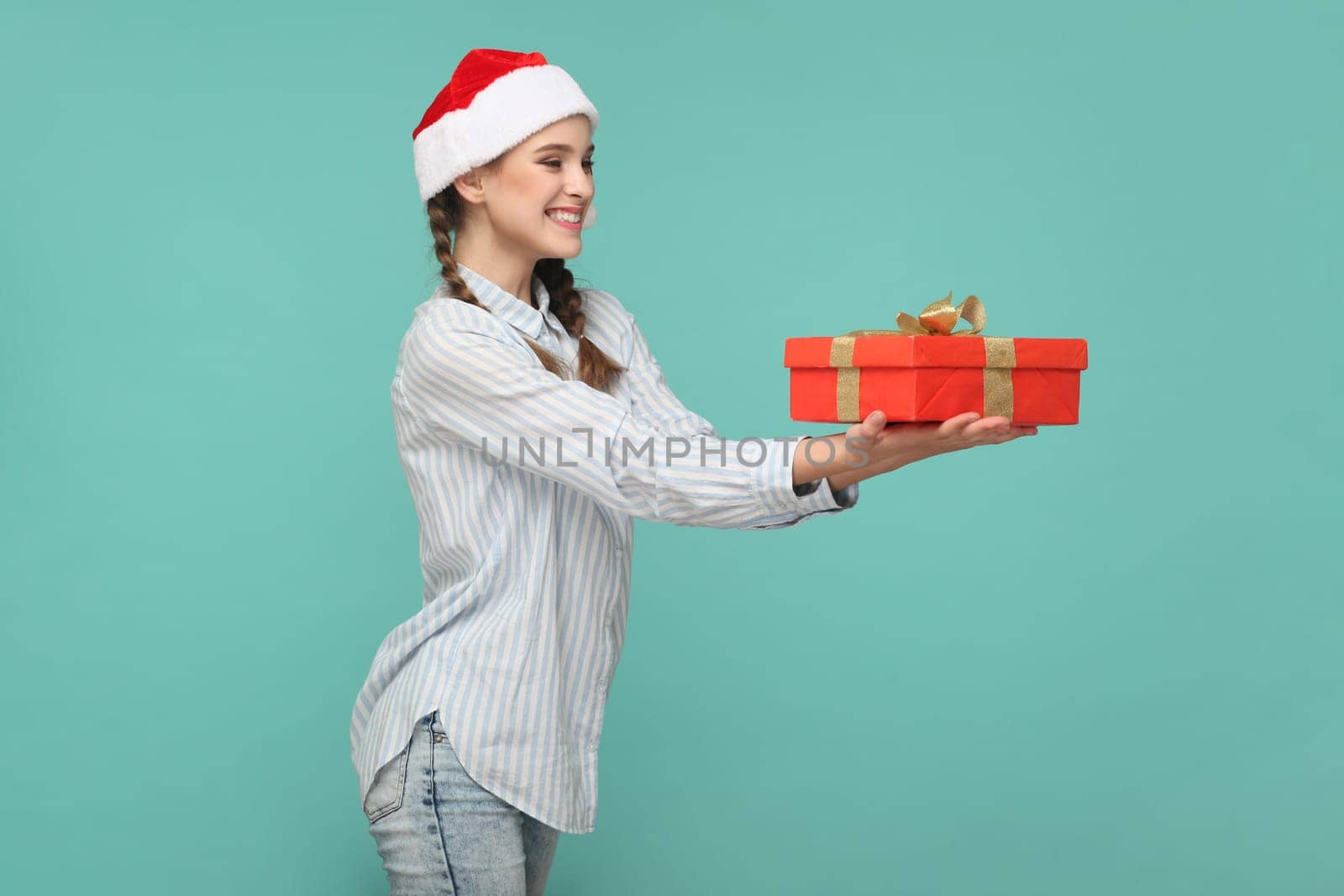 Side view portrait of smiling delighted teenager girl wearing striped shirt and Santa Claus hat, congratulating with new year, giving present. Indoor studio shot isolated on green background.
