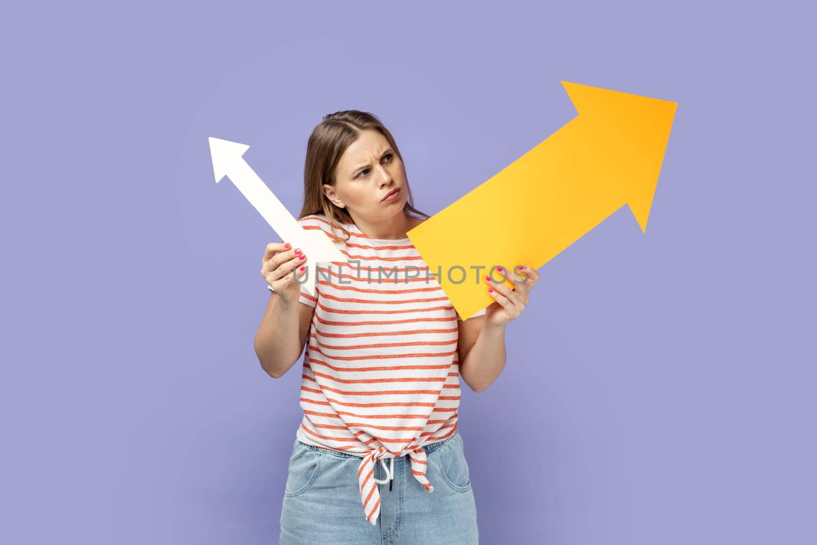 Portrait of confused pensive woman wearing striped T-shirt holding two arrows indicating to different sizes, looking at indicators with puzzlement. Indoor studio shot isolated on purple background.