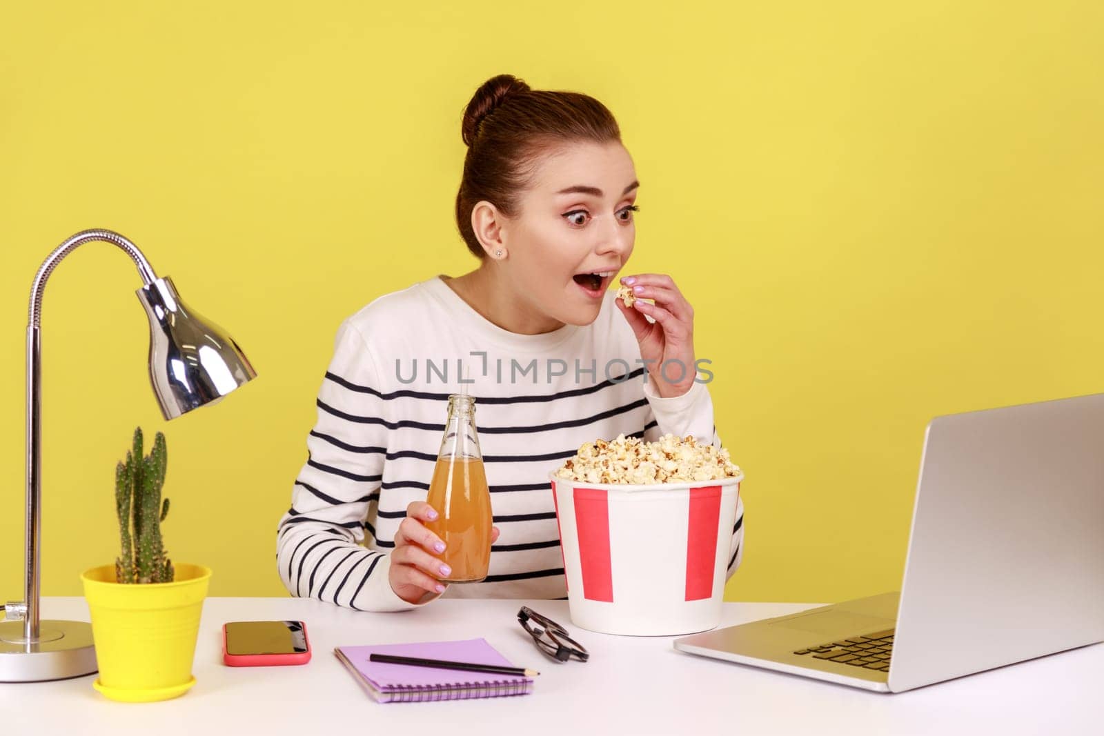 Happy excited woman sitting at workplace and watching movie during resting, eating popcorn, looking at laptop screen with open mouth. Indoor studio studio shot isolated on yellow background.