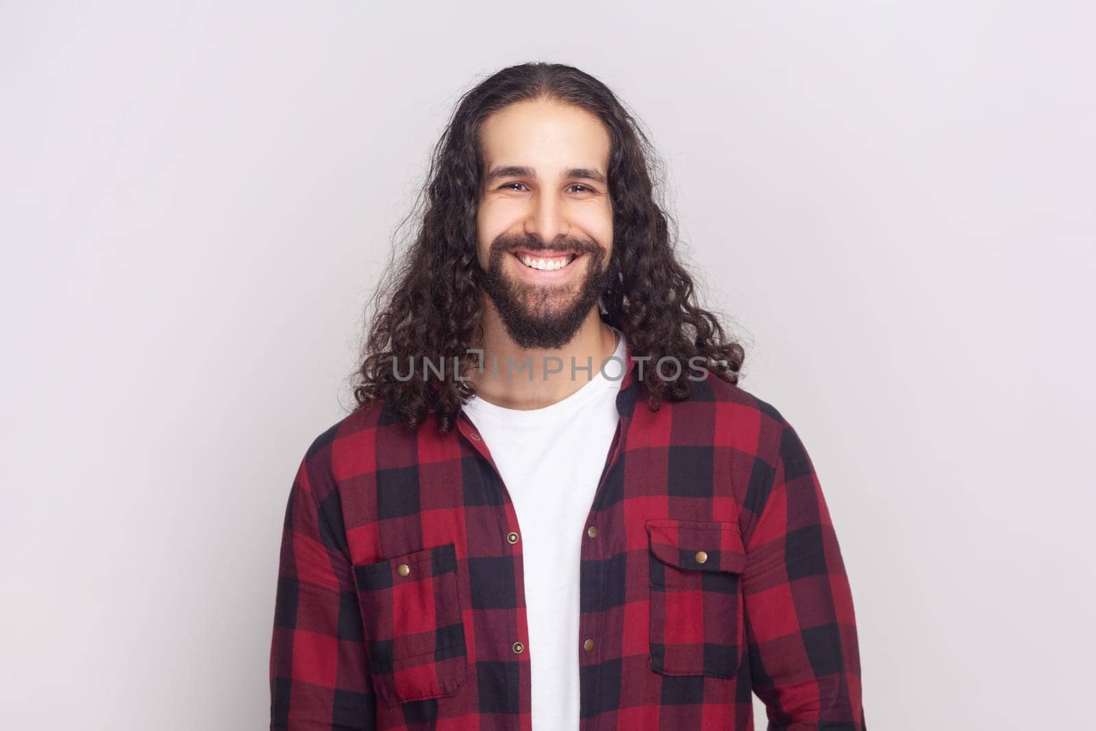 Portrait of satisfied cheerful handsome bearded man with long curly hair in checkered red shirt looks at camera, glad to find suitable well paid job. Indoor studio shot isolated on gray background.