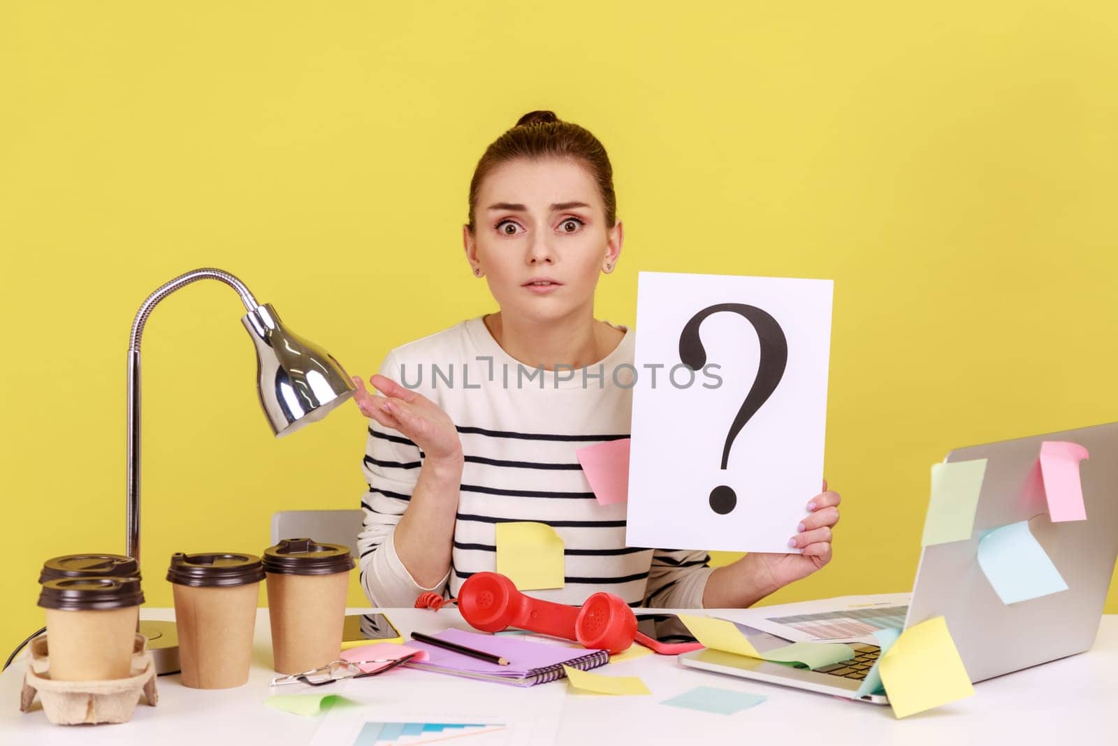 Portrait of shocked astonished woman looking at camera, holding paper with question mark, thinks about tasks, covered with sticky notes. Indoor studio studio shot isolated on yellow background.