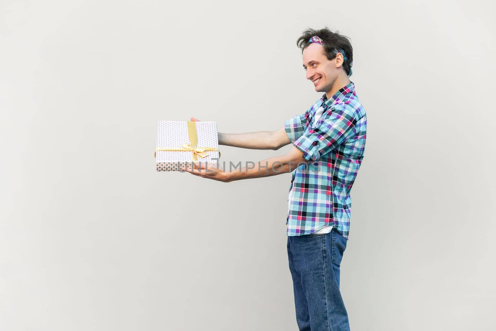 Side view of positive happy smiling standing with outstretched hands, giving present to his girlfriend, wearing blue checkered shirt and headband. Indoor studio shot isolated on gray background.