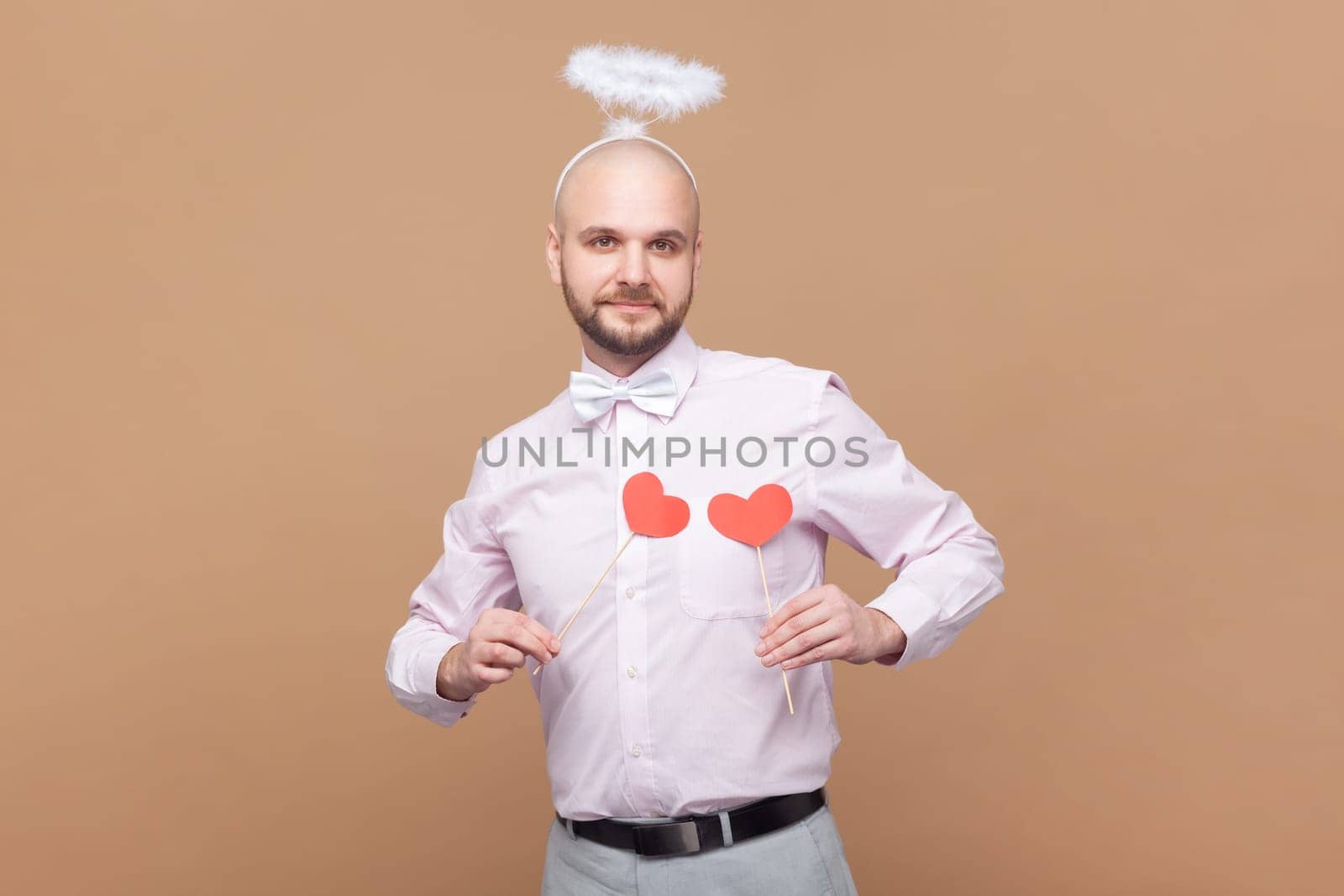 Portrait of cute friendly bald bearded man with nimb over head, holding little red hearts in front of his chest, wearing light pink shirt and bow tie. Indoor studio shot isolated on brown background.