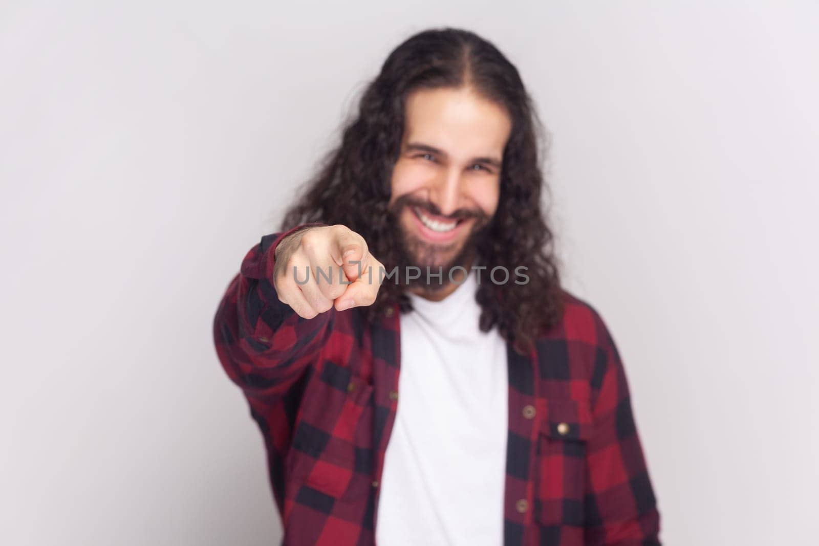 You got it. Happy bearded man with long curly hair in checkered red shirt points at camera with index fingers, picks someone smiles broadly. Indoor studio shot isolated on gray background.