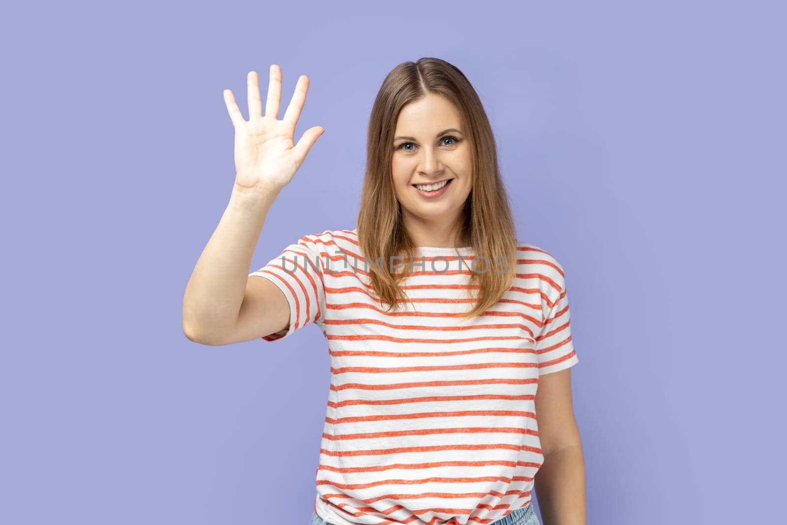 Portrait of friendly positive blond woman wearing striped T-shirt smiles toothily, raises palm greets friend, saying hello or goodbye. Indoor studio shot isolated on purple background.