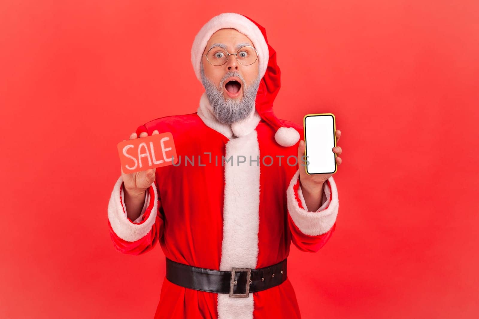 Portrait of shocked elderly man with gray beard wearing santa claus costume holding smart phone with empty screen and sale card, huge discounts. by Khosro1