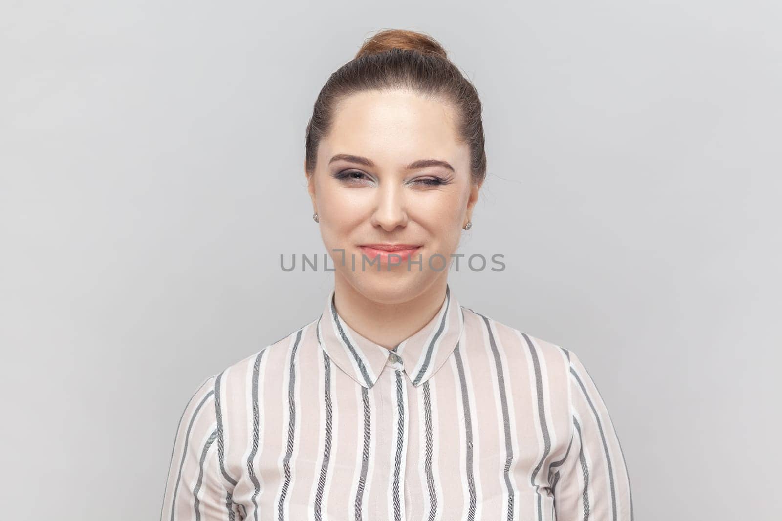 Portrait of beautiful playful woman wearing striped shirt standing looking at camera and winking, flirting with her boyfriend, smiling happily. Indoor studio shot isolated on gray background.