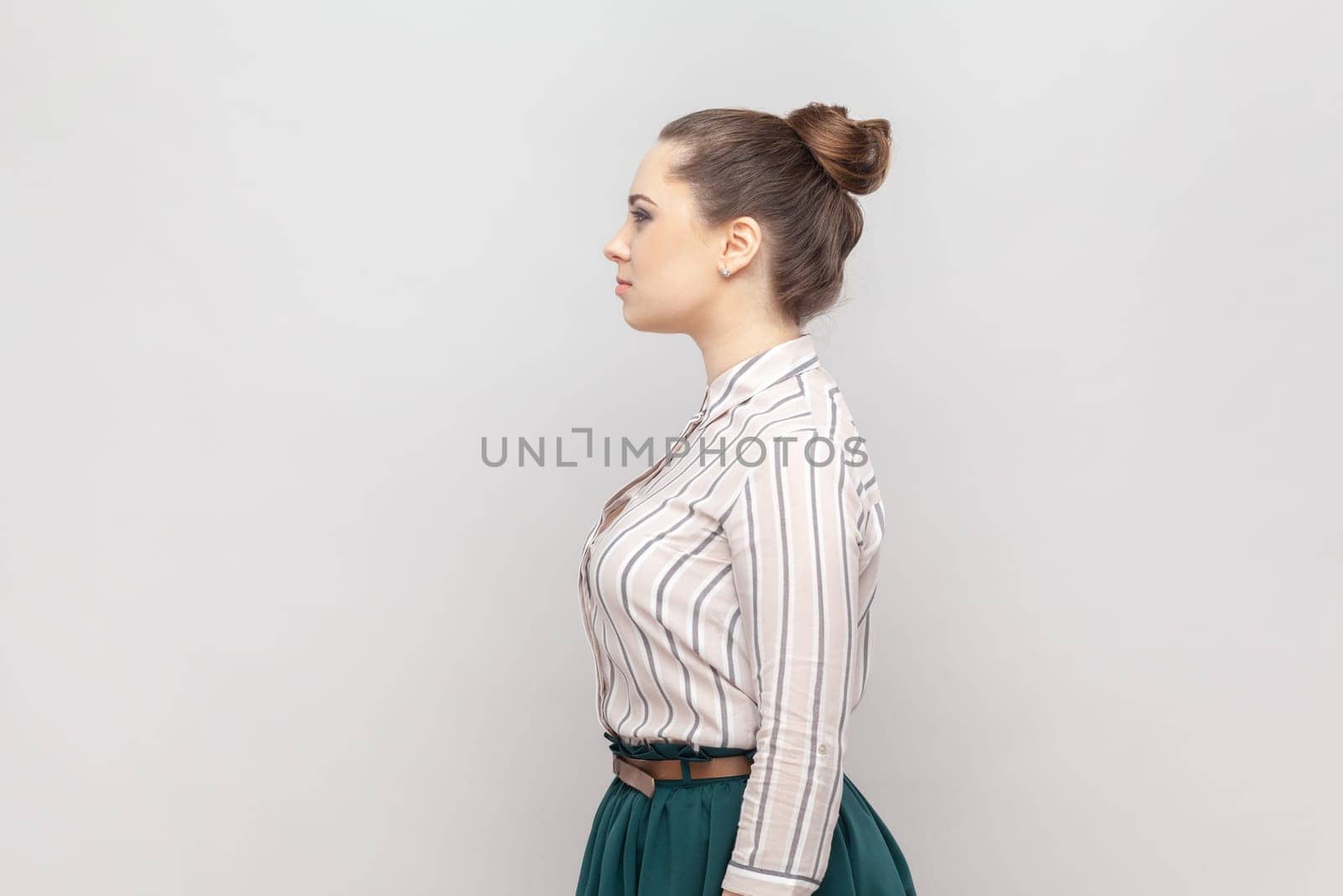 Side view portrait of serious strict woman with bun hairstyle wearing striped shirt and green skirt standing looking ahead, having bossy expression. Indoor studio shot isolated on gray background.