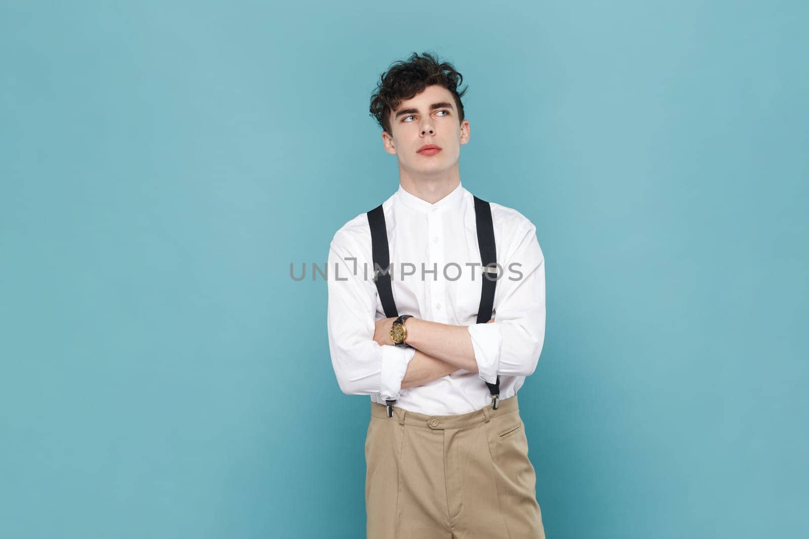 Portrait of handsome pensive thoughtful man wearing white shirt and suspender crossing his hands and looking away, thinking. Indoor studio shot isolated on blue background.