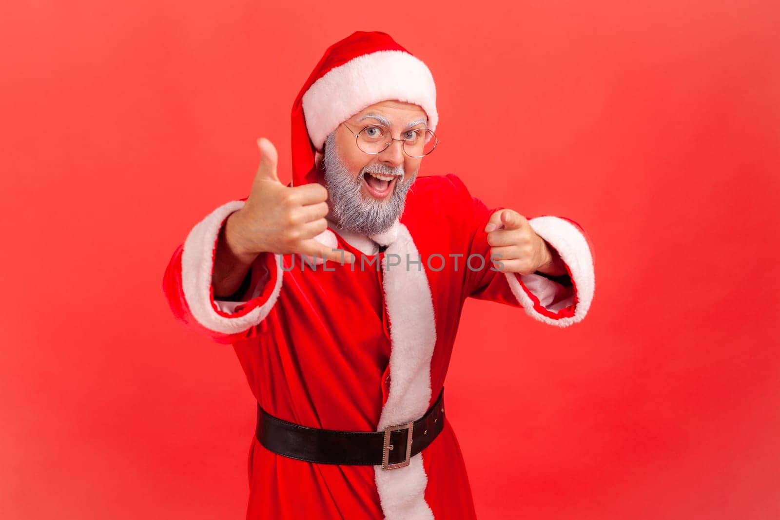 Elderly man with gray beard wearing santa claus costume holding fingers near ear showing telephone gesture, pointing at camera, waiting for your call. by Khosro1
