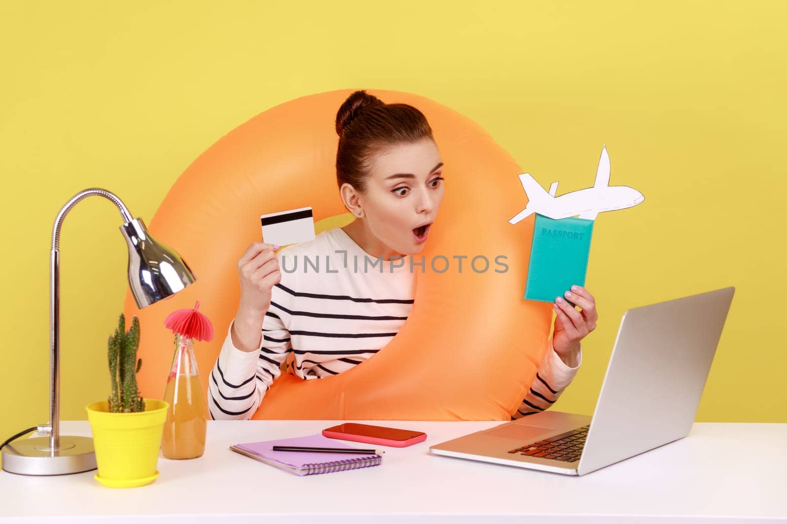 Woman with rubber ring showing credit card and passport, looking at laptop display with open mouth. by Khosro1