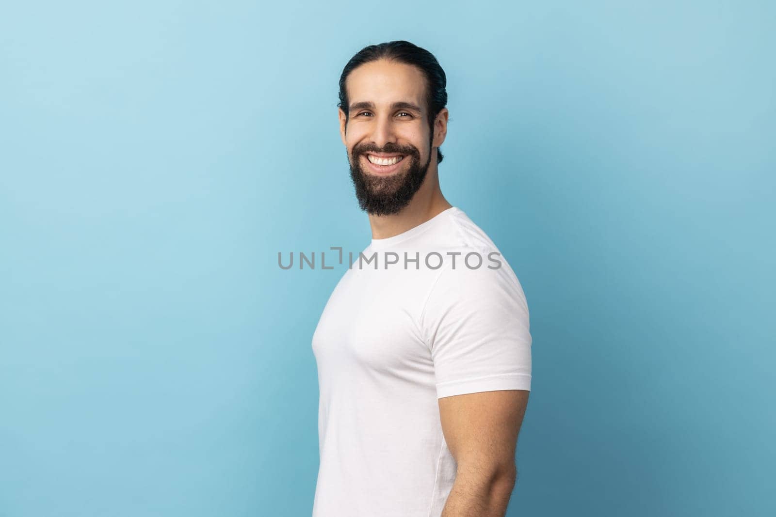 Side view of bearded handsome man wearing white T-shirt standing looking at camera with satisfied face and smiling, expressing happiness. Indoor studio shot isolated on blue background.