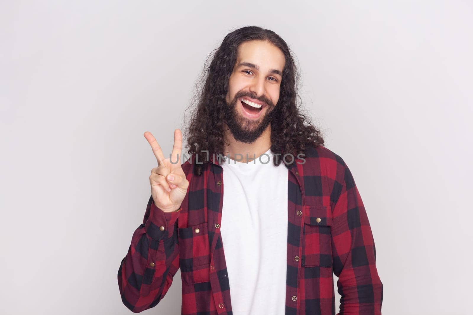 Portrait of optimistic glad bearded man with long curly hair in checkered red shirt with positive facial expression, shows V sign or victory gesture. Indoor studio shot isolated on gray background.
