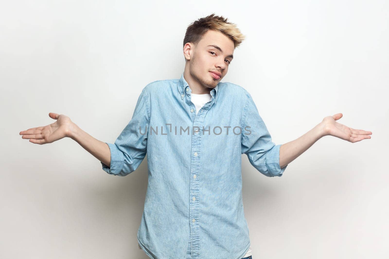 Portrait of doubtful puzzled attractive man wearing denim shirt standing with spread hands, looking at camera and shrugging shoulders. Indoor studio shot isolated on gray background.