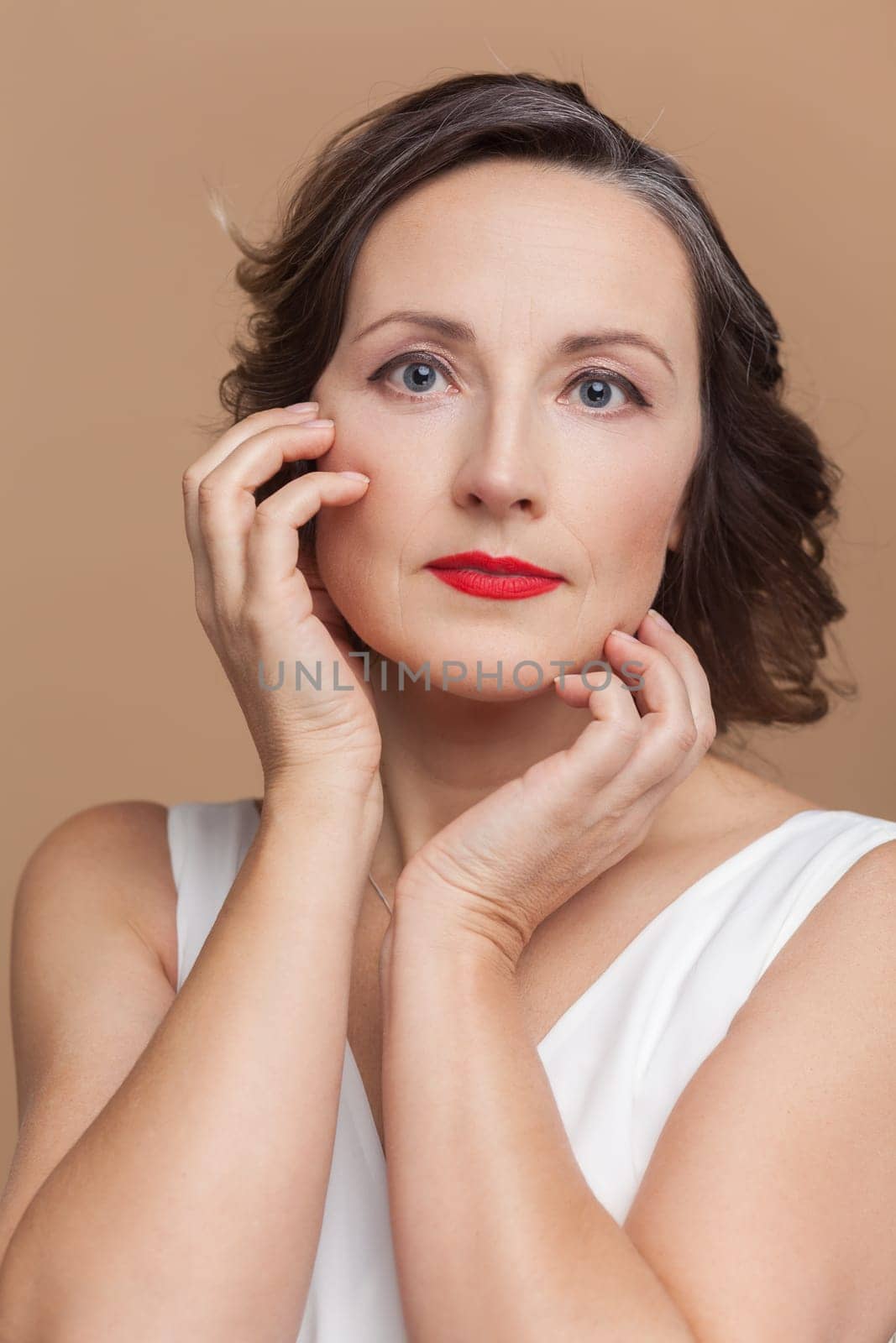 Closeup portrait of calm attractive beautiful middle aged woman with wavy hair and makeup, looking at camera, wearing white dress. Indoor studio shot isolated on light brown background.