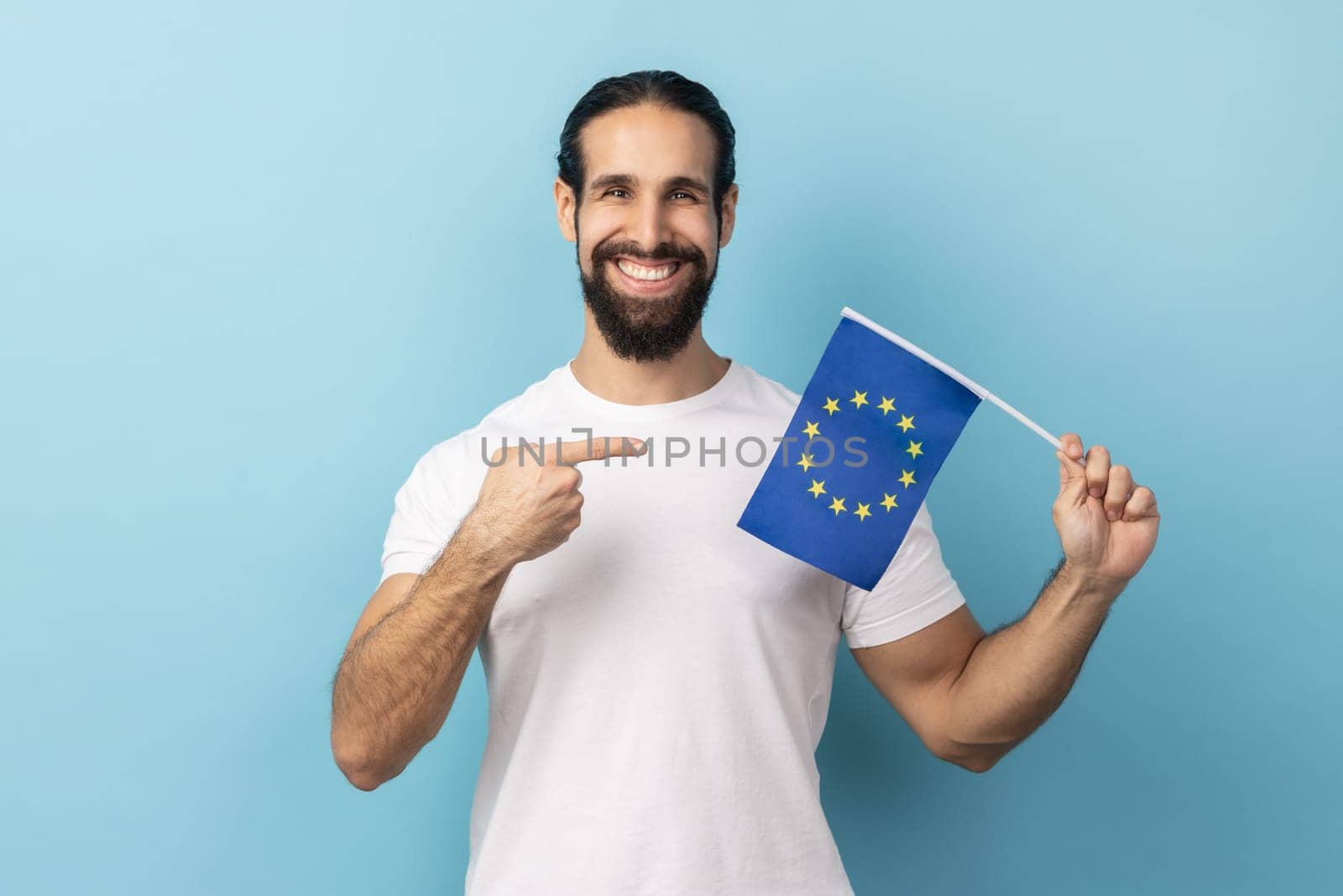 Portrait of man with beard wearing white T-shirt smiling broadly and pointing flag of European Union, symbol of Europe, EU association and community. Indoor studio shot isolated on blue background.