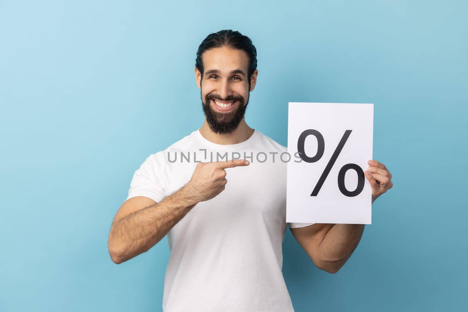 Portrait of attractive optimistic man with beard wearing white T-shirt holding and pointing paper with percent sign inscription, looking at camera. Indoor studio shot isolated on blue background.