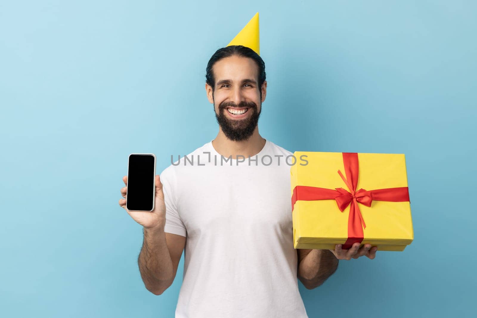Portrait of man with beard wearing white T-shirt and in party cone holding in hands yellow wrapped present box and smart phone with empty display. Indoor studio shot isolated on blue background.
