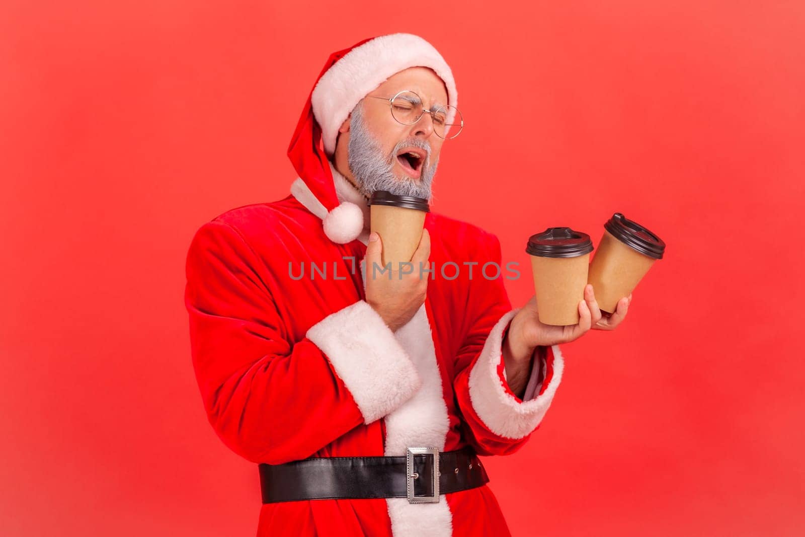 Tired sleepy elderly man with gray beard wearing santa claus costume holding many paper disposable cups with coffee, needs much energy. Indoor studio shot isolated on red background.